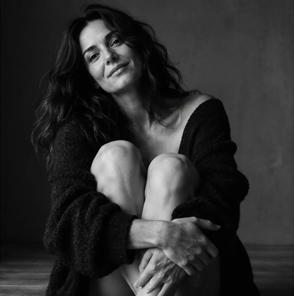 Ritratto intimo in bianco e nero di una donna seduta che abbraccia le ginocchia, con un cardigan scuro e un sorriso